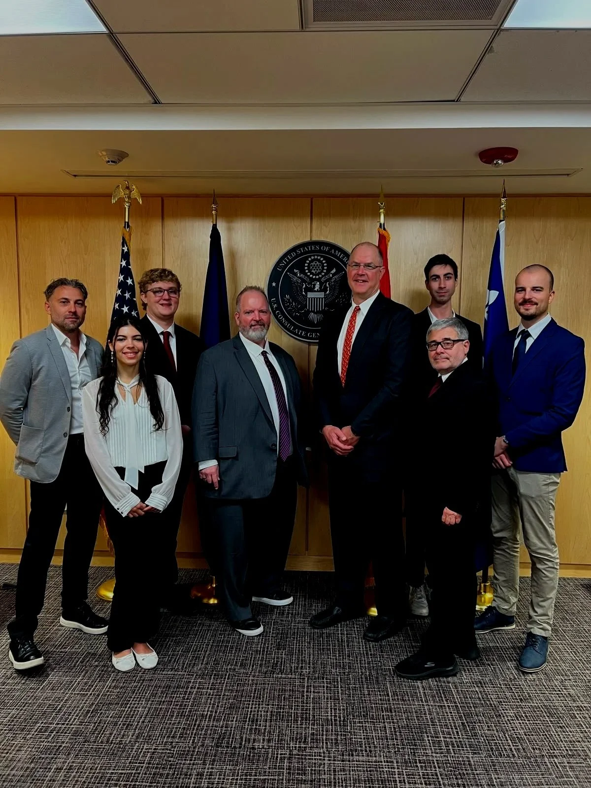 Group of nine people in business attire posing for a photo in a formal setting with US flags and a government emblem in the background.