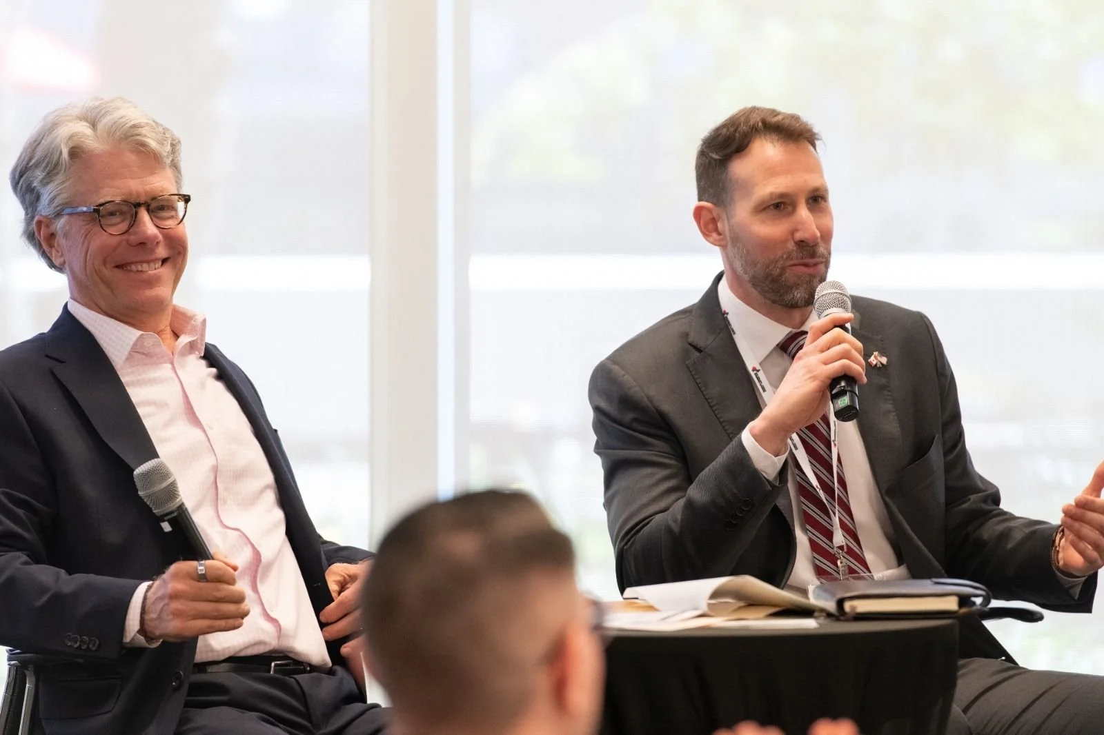 Two men in business suits participating in a panel discussion, one smiling and holding a microphone, the other speaking and holding a microphone.