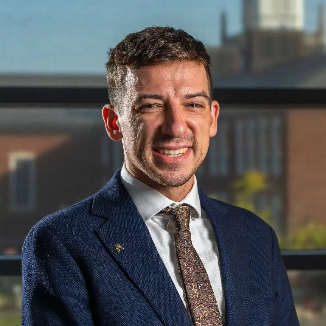 Portrait of a smiling man in a navy suit, white shirt, and patterned tie, standing in front of a window with a cityscape background.