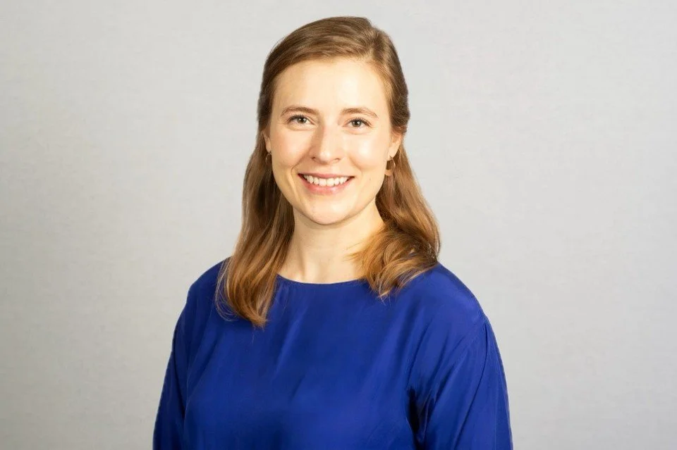 A woman with shoulder-length light brown hair wearing a blue blouse, smiling at the camera against a plain gray background.
