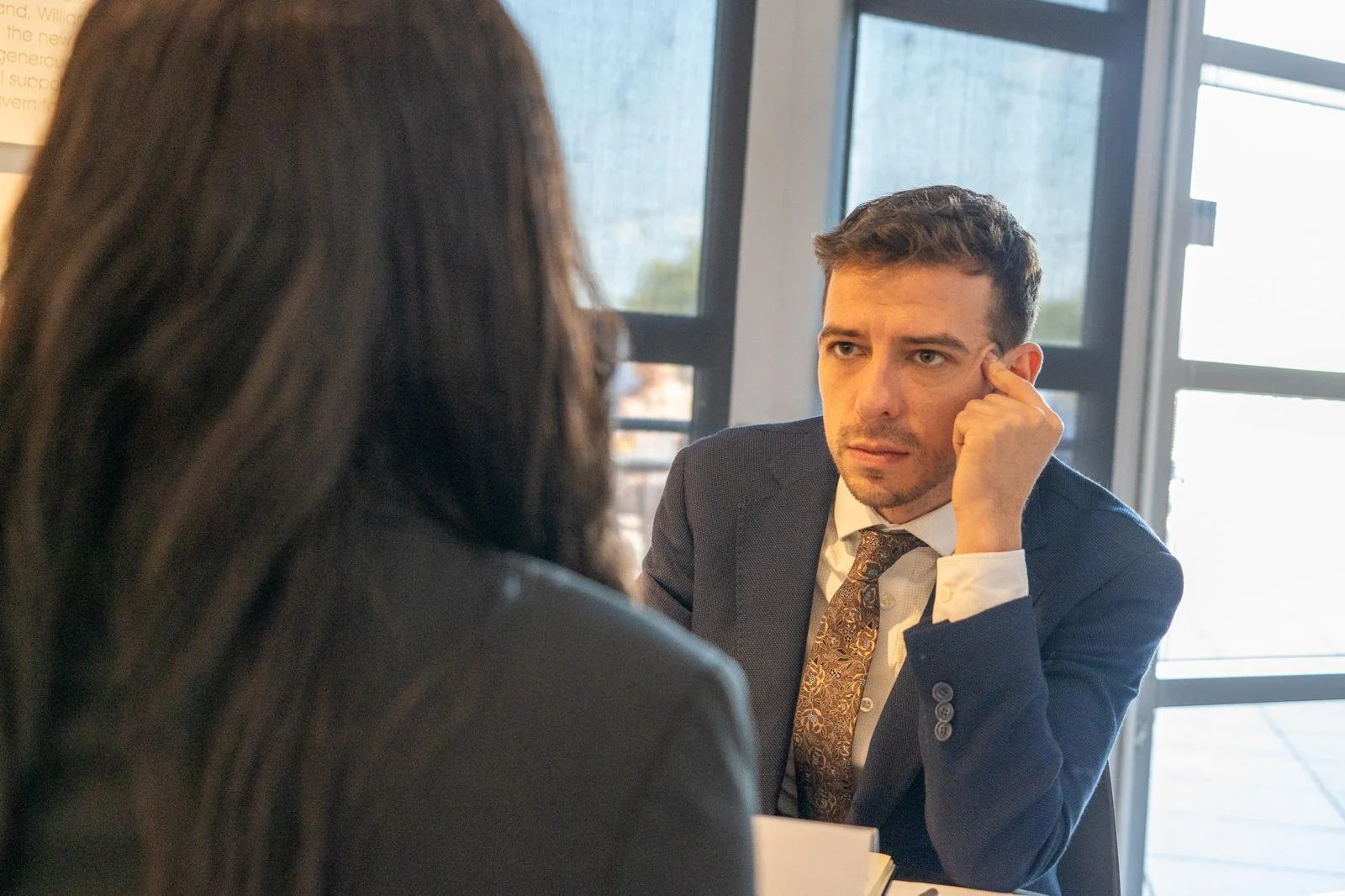 A man in a business suit talking to a woman with dark hair during an interview or meeting in a modern office.