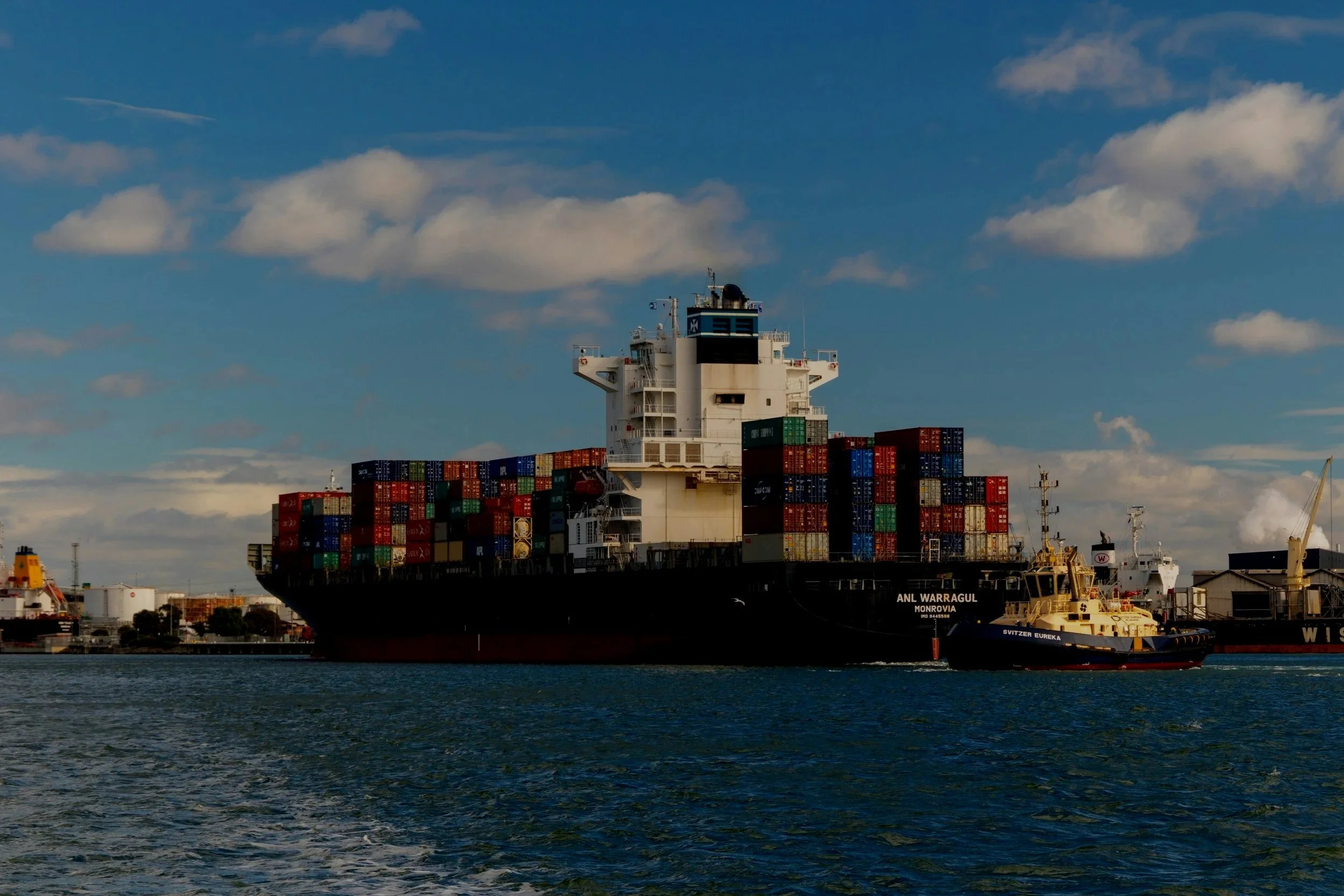 Large cargo ship named 'ANL Warragul' carrying multicolored shipping containers on the water with a blue sky and white clouds overhead.