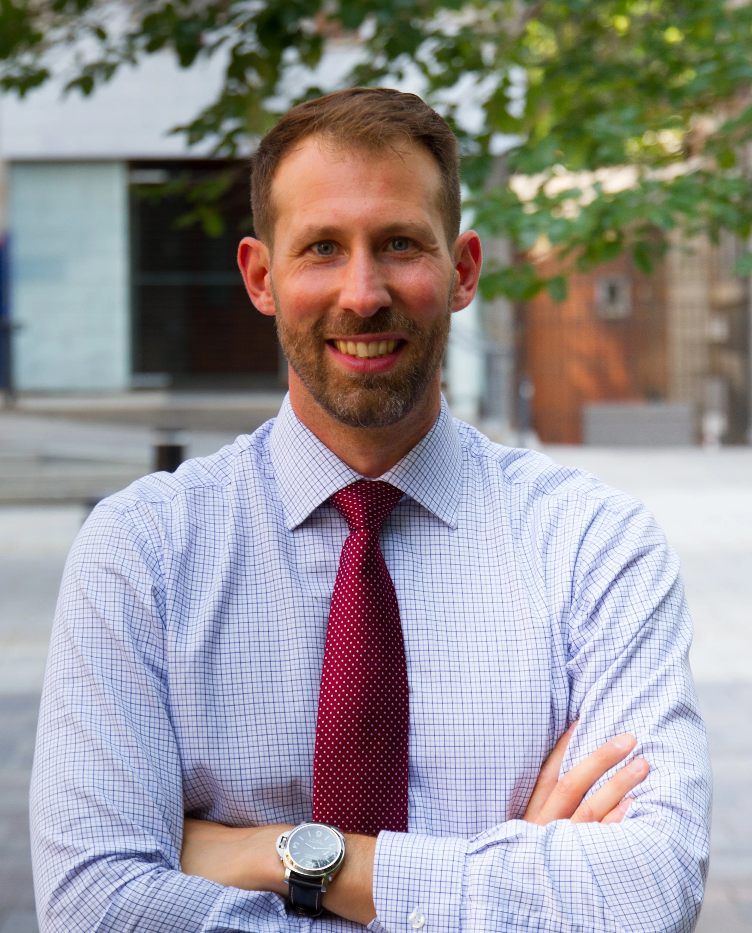 Close-up of a man smiling outdoors, wearing a checkered dress shirt, red tie, and wristwatch, with arms crossed and background of green leaves and buildings.