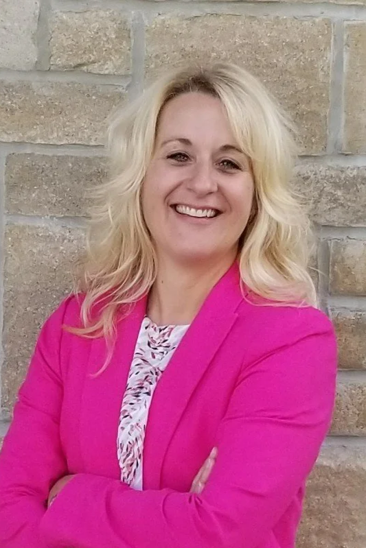 A woman with blonde hair, smiling, wearing a bright pink blazer and a patterned blouse, standing in front of a beige stone wall.