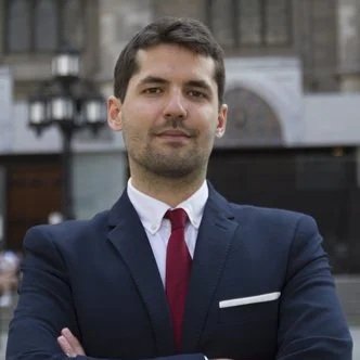 A man in a dark suit with a white shirt and maroon tie standing outdoors with a serious expression.