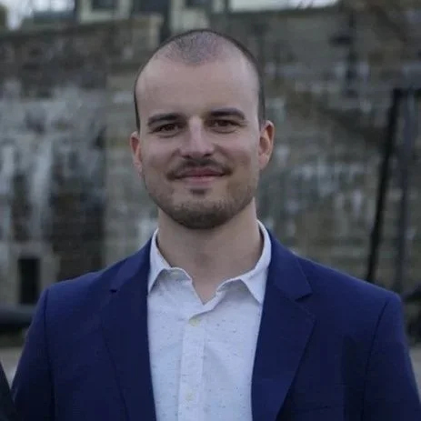 Portrait of a young man with a beard and short hair, wearing a dark blue suit jacket and white shirt, standing outdoors with a blurred background.