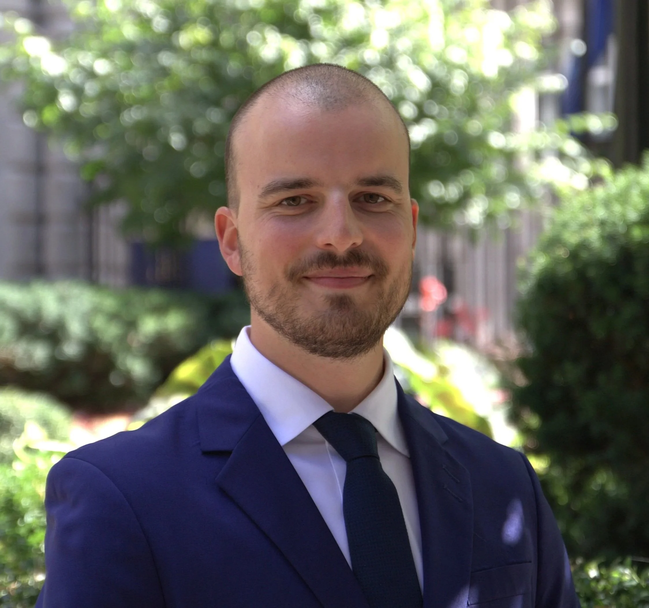 Portrait of a young man with a beard and short hair, wearing a dark blue suit jacket and white shirt, standing outdoors with a blurred background.