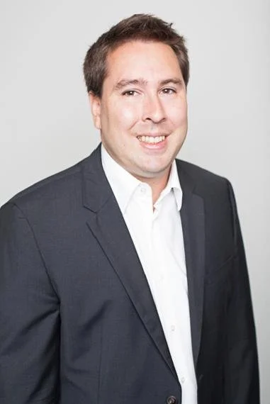 Professional man in a dark suit and white shirt smiling against a plain background.