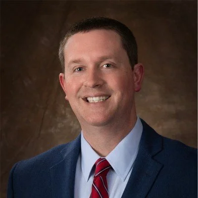A professional headshot of a man in a suit and tie smiling against a brown background.