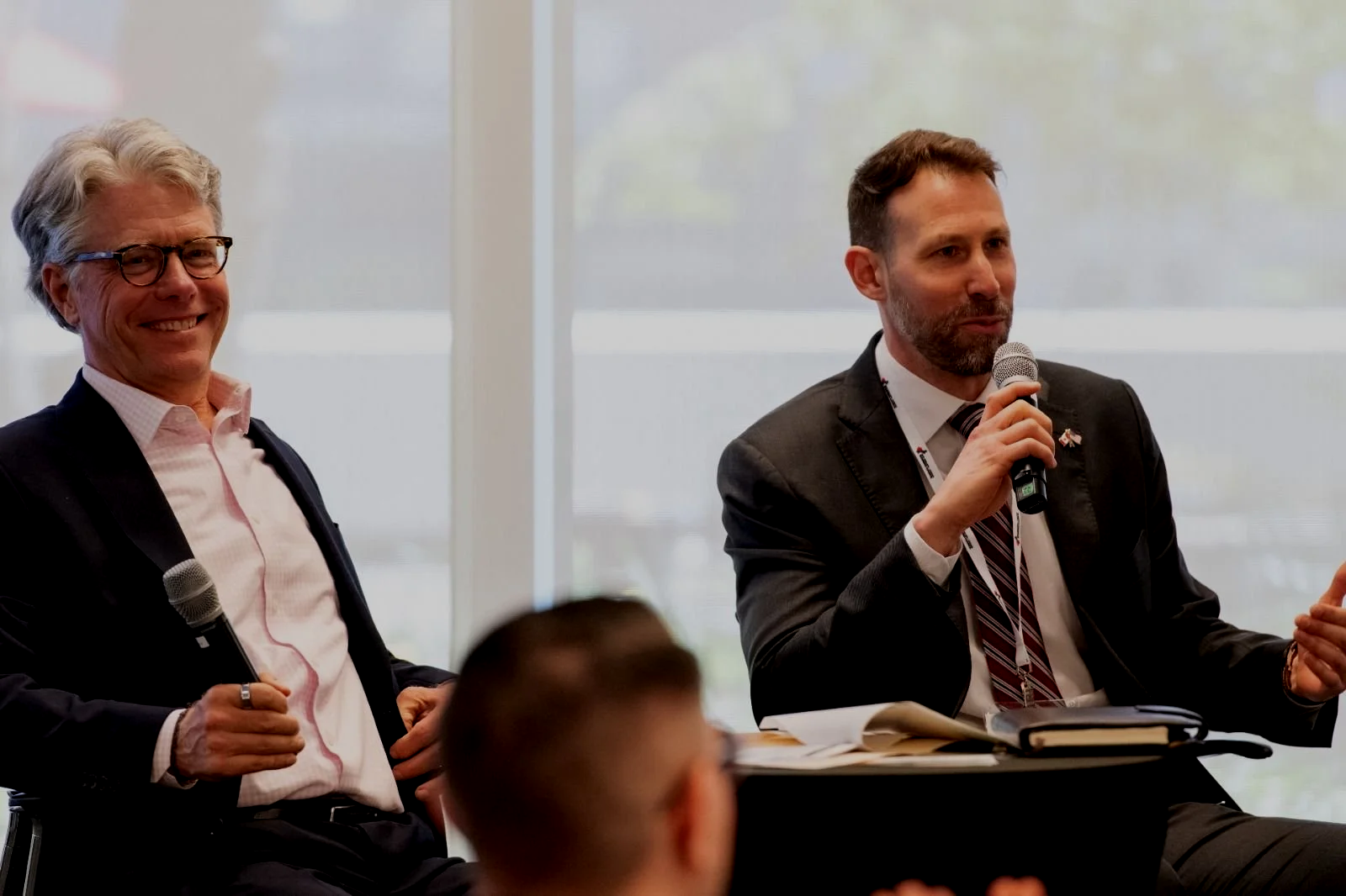 Two men sitting at a panel discussion, one smiling with glasses and holding a microphone, the other speaking into a microphone while wearing a dark suit and striped tie, in front of a window.