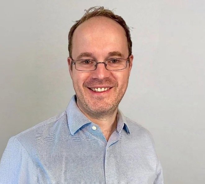 A portrait of a smiling man with glasses, wearing a light blue button-up shirt, against a plain light-colored background.