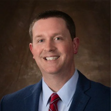 Professional portrait of a smiling man with short brown hair wearing a dark blue suit, white shirt, and red striped tie, against a brown background.