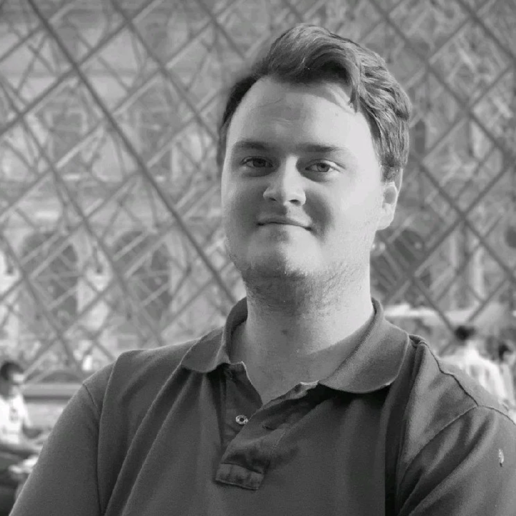 A young man with short hair, wearing a polo shirt and a backpack, stands indoors in front of a geometric patterned wall, smiling slightly.