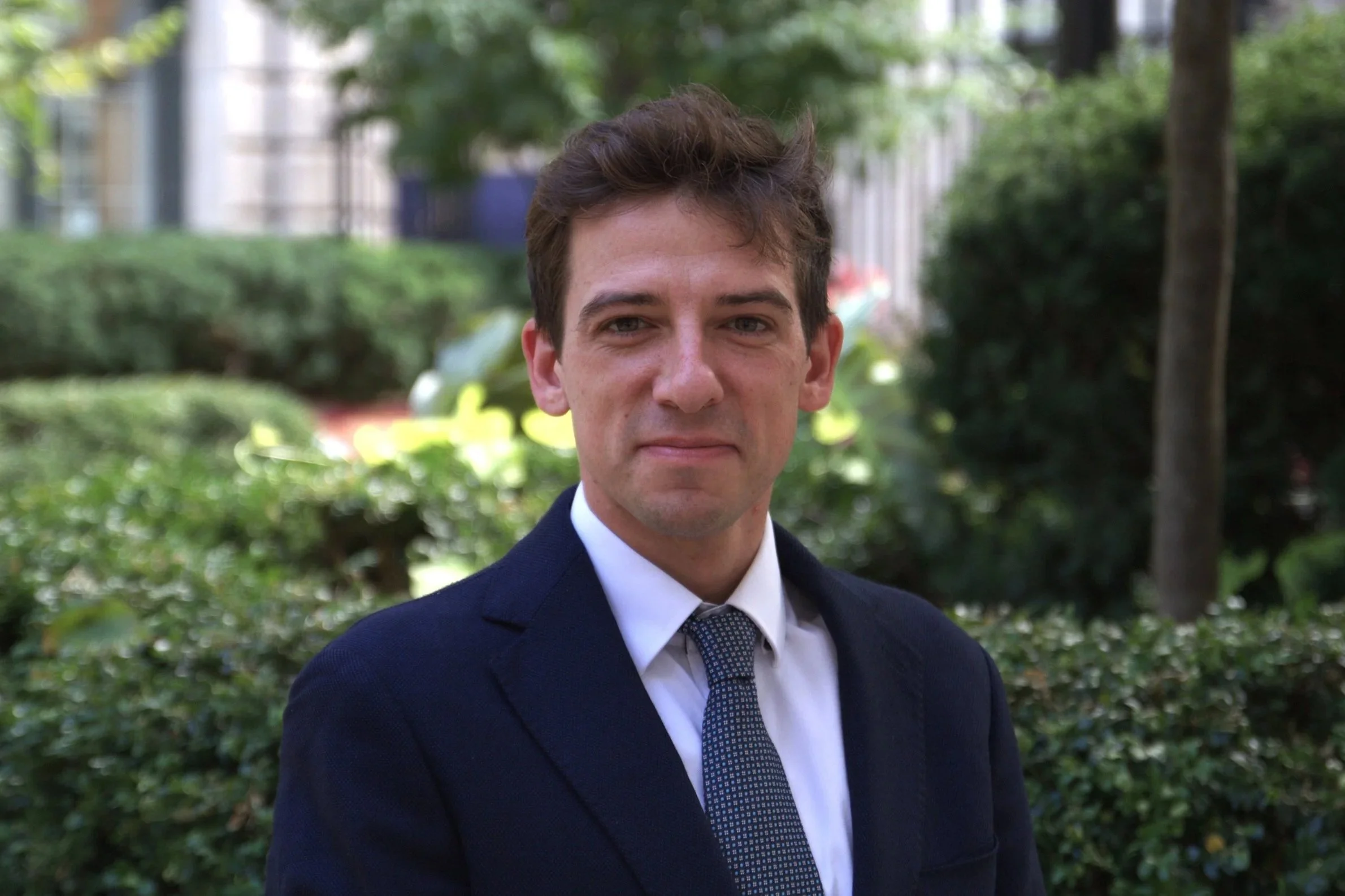 Portrait of a smiling man in a navy suit, white shirt, and patterned tie, standing in front of a window with a cityscape background.