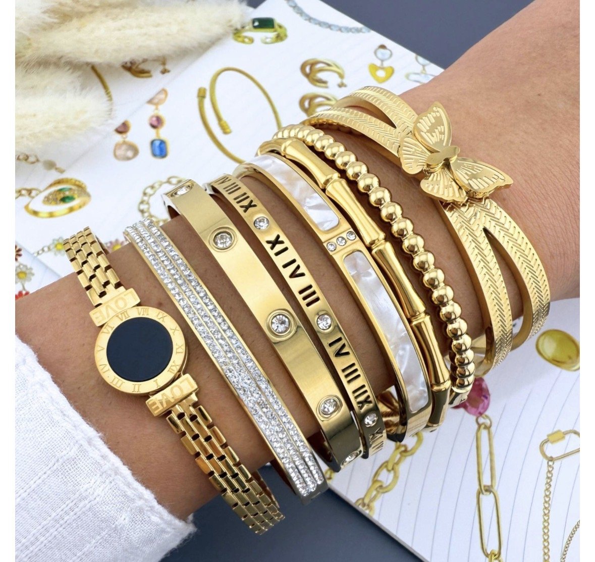 A person's wrist adorned with multiple gold and silver bracelets, including watches, bangles, and rings, with jewelry catalogs and gold jewelry illustrations in the background.