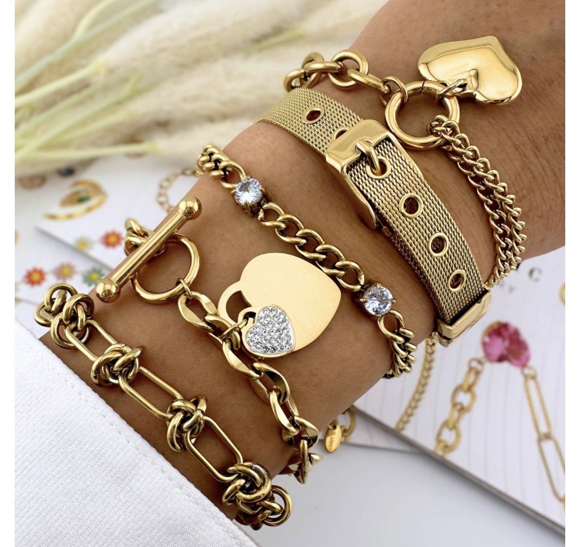 A wrist decorated with multiple gold bracelets, including a chain link bracelet, a leather strap bracelet with holes, and a bracelet with a heart-shaped charm, some adorned with small clear stones.