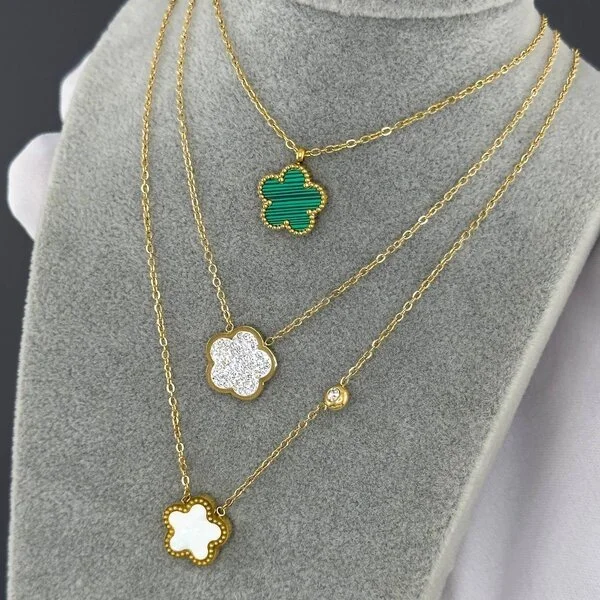 Four gold layered necklaces with clover-shaped pendants displayed on a gray jewelry stand. The top pendant is green, the second is encrusted with white diamonds, the third has a small single diamond, and the bottom pendant is white.