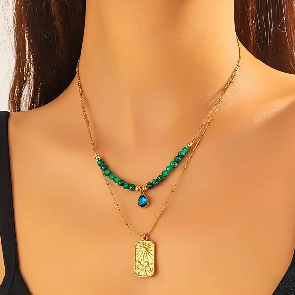 Close-up of a woman's neck wearing two layered gold necklaces, one with green beads and a blue pendant, the other with a gold rectangular pendant with floral engravings.