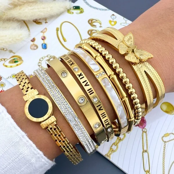 Multiple gold and silver jewelry bracelets on a person's wrist, including watches, bangles with Roman numerals, a butterfly charm, and a beaded bracelet, with jewelry catalog pages in the background.