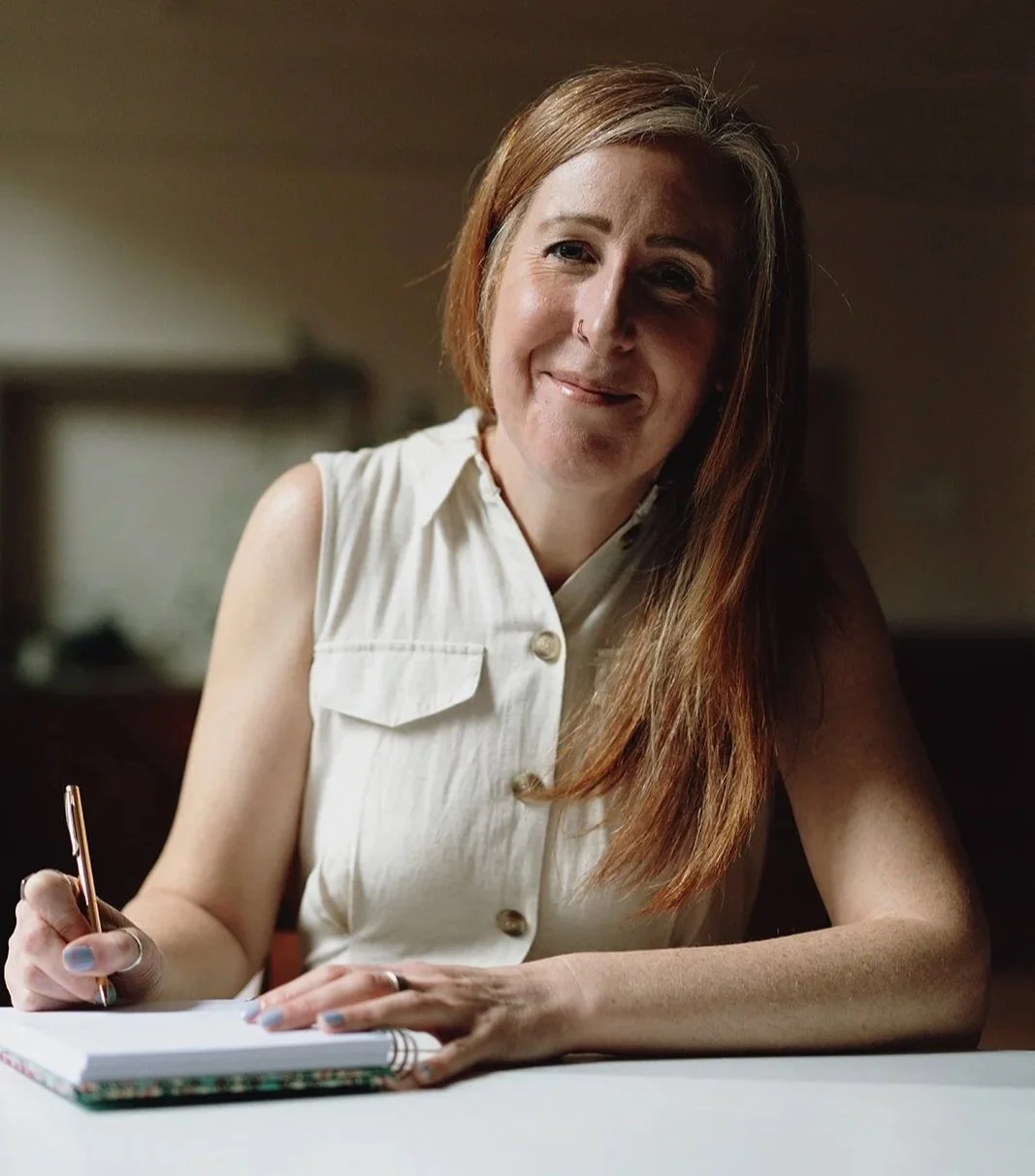 A woman with long red hair and a nose ring sitting at a table, smiling, holding a pen in her right hand, with a notebook or journal open in front of her.