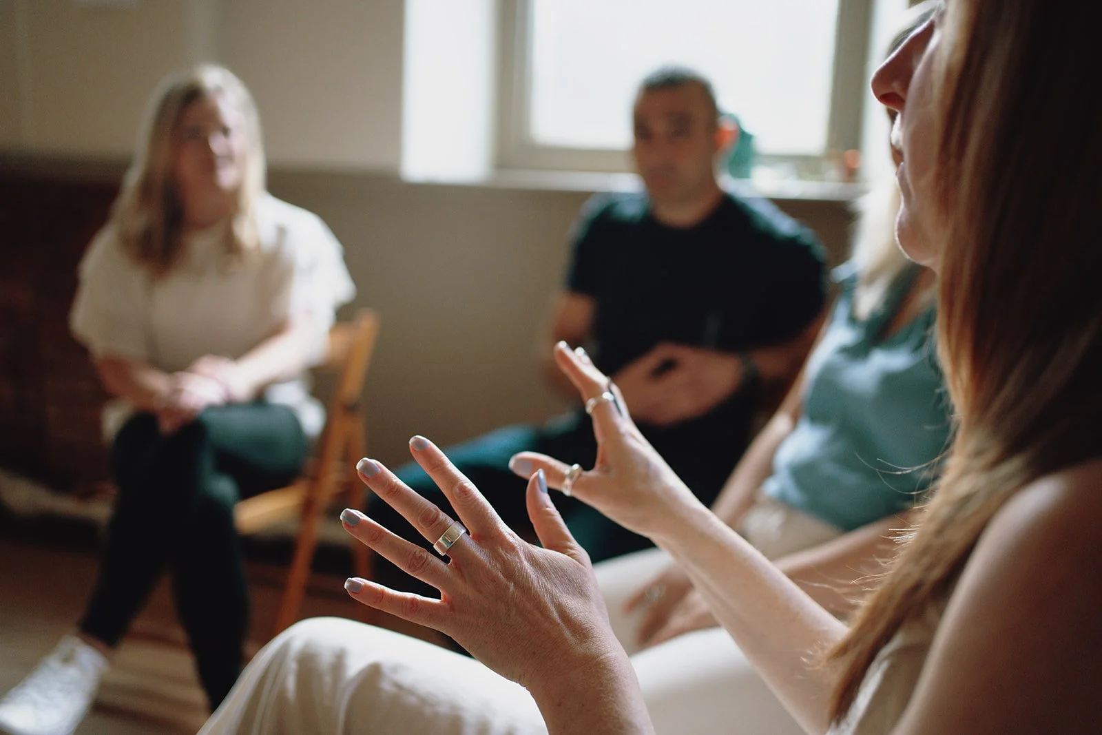 A group of four people engaged in a conversation in a room with natural lighting, focus on a woman gesturing with her hands in the foreground.