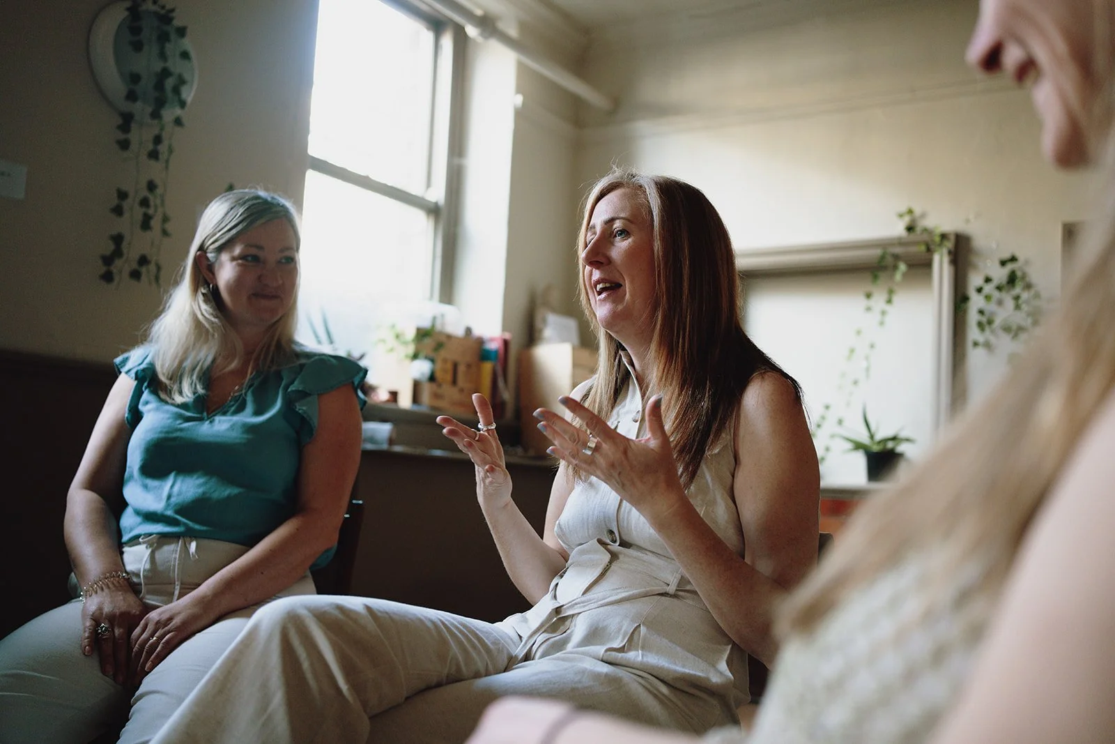 Three women sitting in a room, engaged in conversation. The woman in the center with brown hair is speaking, while the other two women, one with blonde hair and the other partially visible, listen. The room has a window with natural light, some plants, and decorative items.