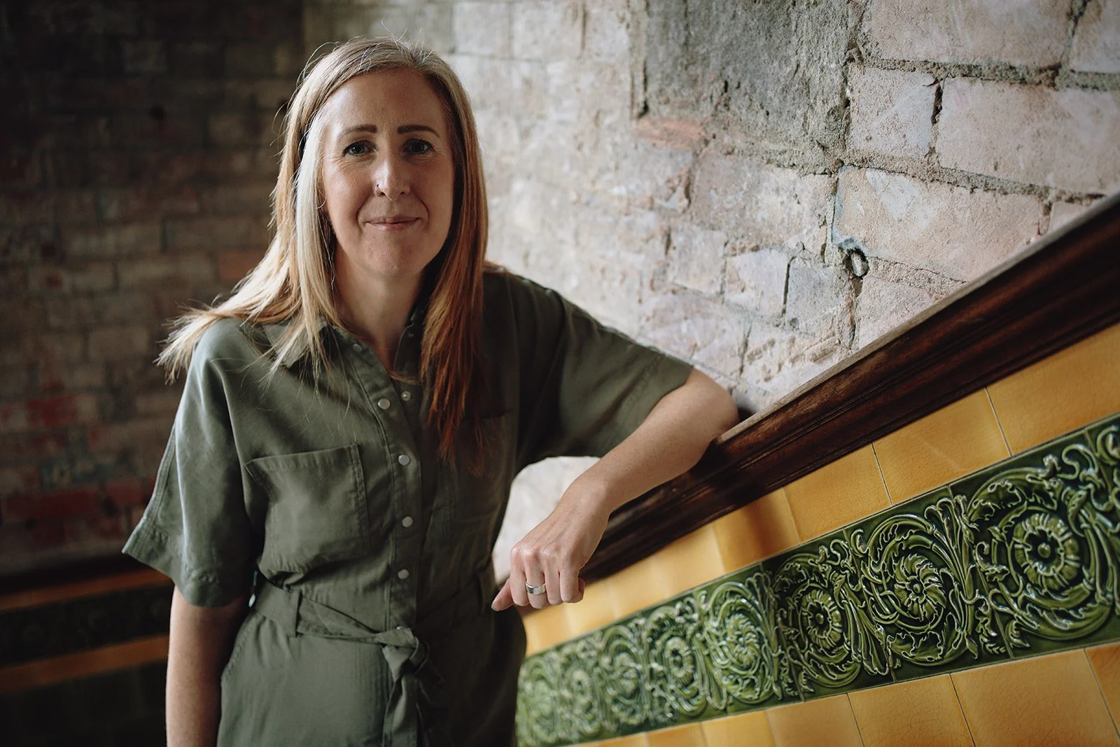 A woman with shoulder-length light brown hair, wearing a green button-up shirt, leaning on a decorative green and yellow tiled surface against a brick wall.