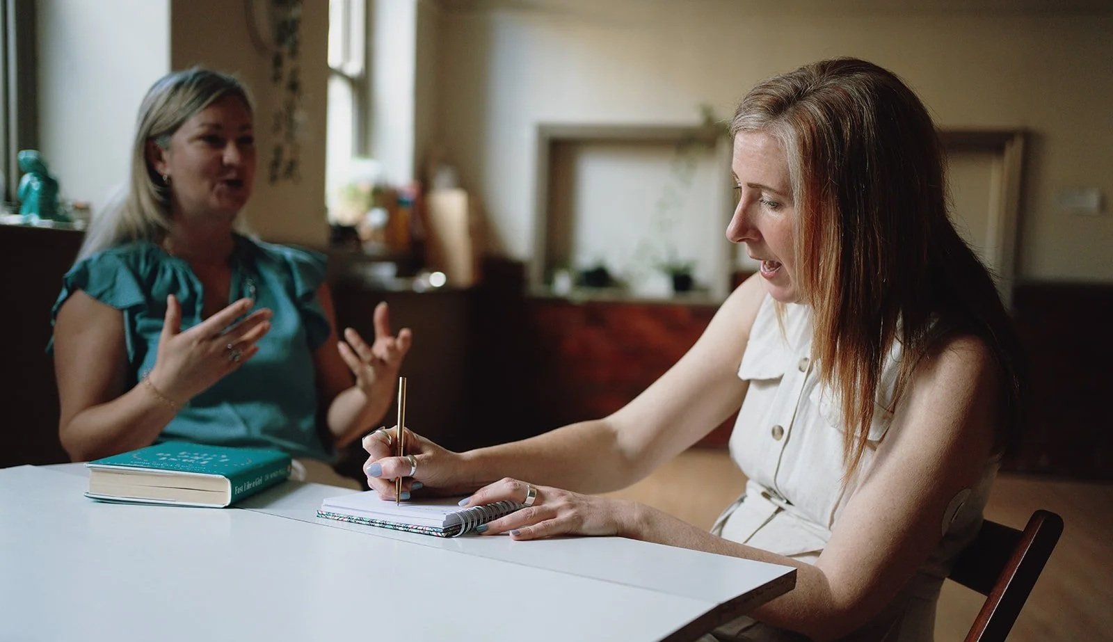 Two women sitting at a white table, engaged in a conversation. One woman is taking notes or signing a document while the other woman is gesturing with her hands, speaking passionately. The room has a window in the background with natural light.