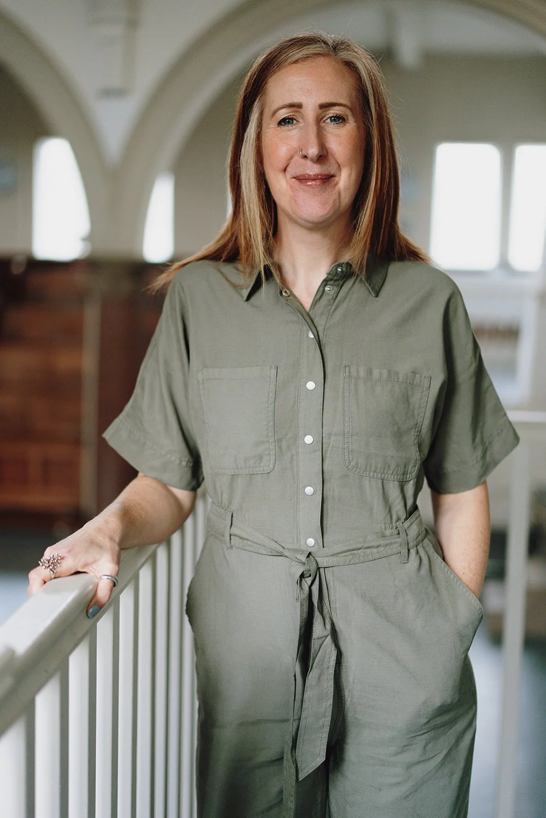 A woman with reddish-brown hair and a nose ring smiling and standing indoors, leaning on a white railing, wearing a green jumpsuit with short sleeves.