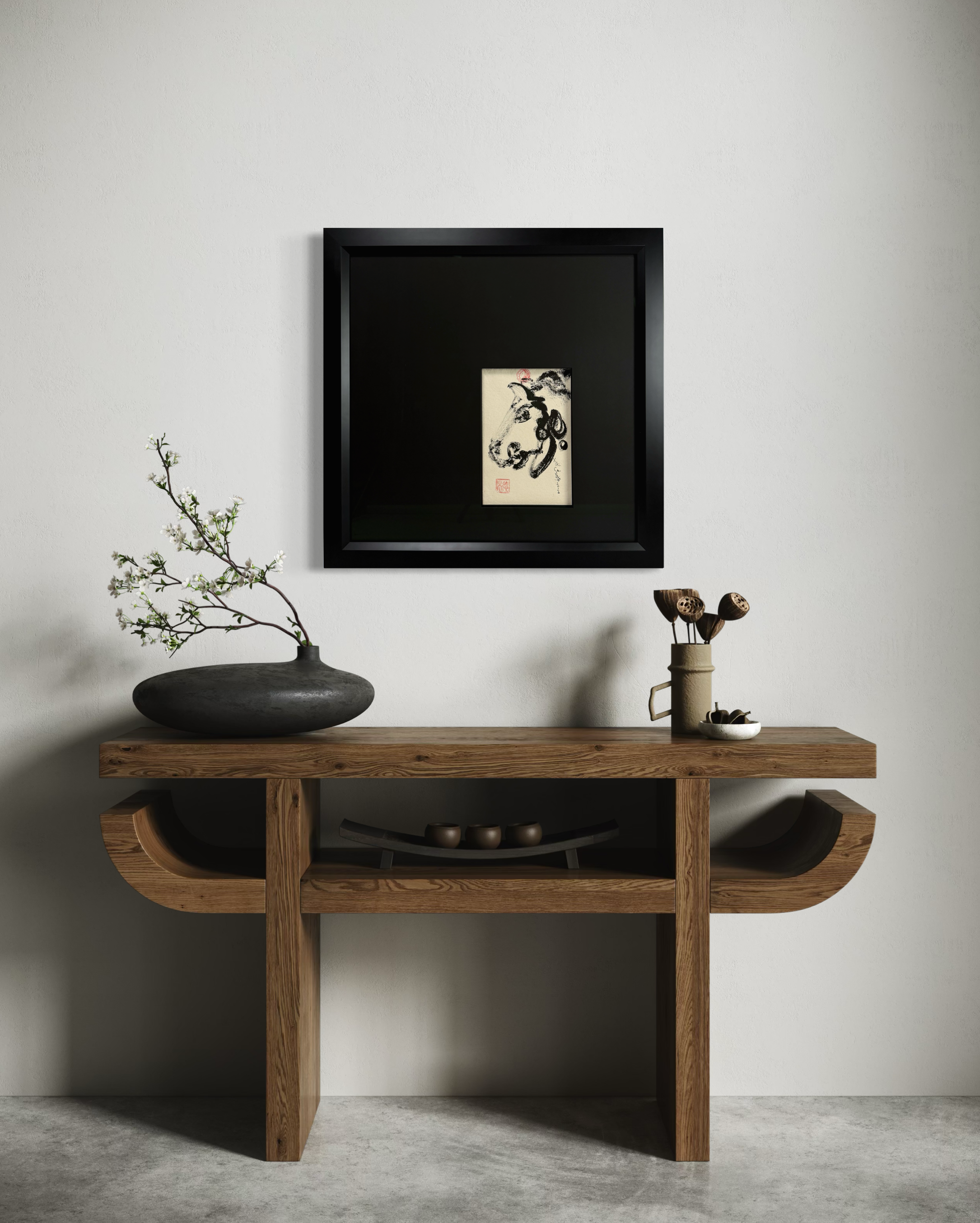 A wooden console table with decorative objects, including a branch with white flowers in a black vase, small bowls, and a ceramic cup, against a plain wall with a framed black-and-white Asian ink painting hung above.