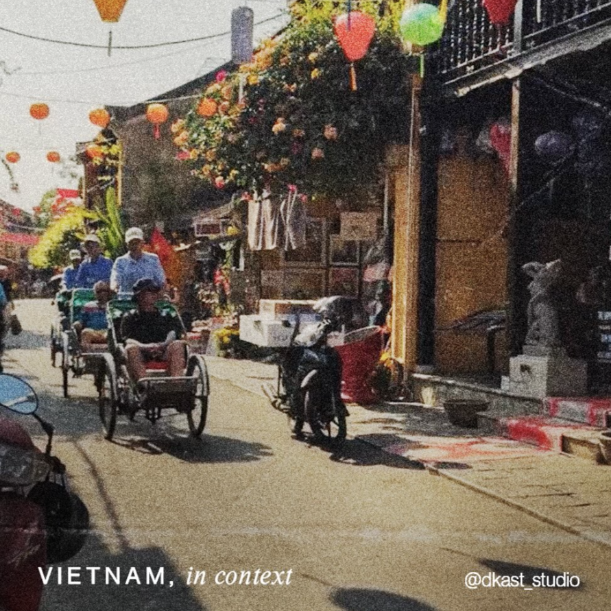 Street scene in Vietnam with rickshaws, motorbikes, and colorful lanterns hanging overhead, featuring shops and a stone statue.
