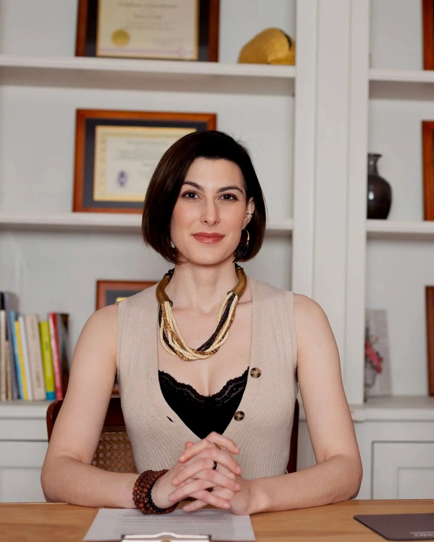 A woman with short dark hair sitting at a wooden table, hands clasped, in front of a beige vest with black lace and gold buttons, wearing a layered necklace and bracelets, with books and framed certificates or diplomas on white shelves behind her.