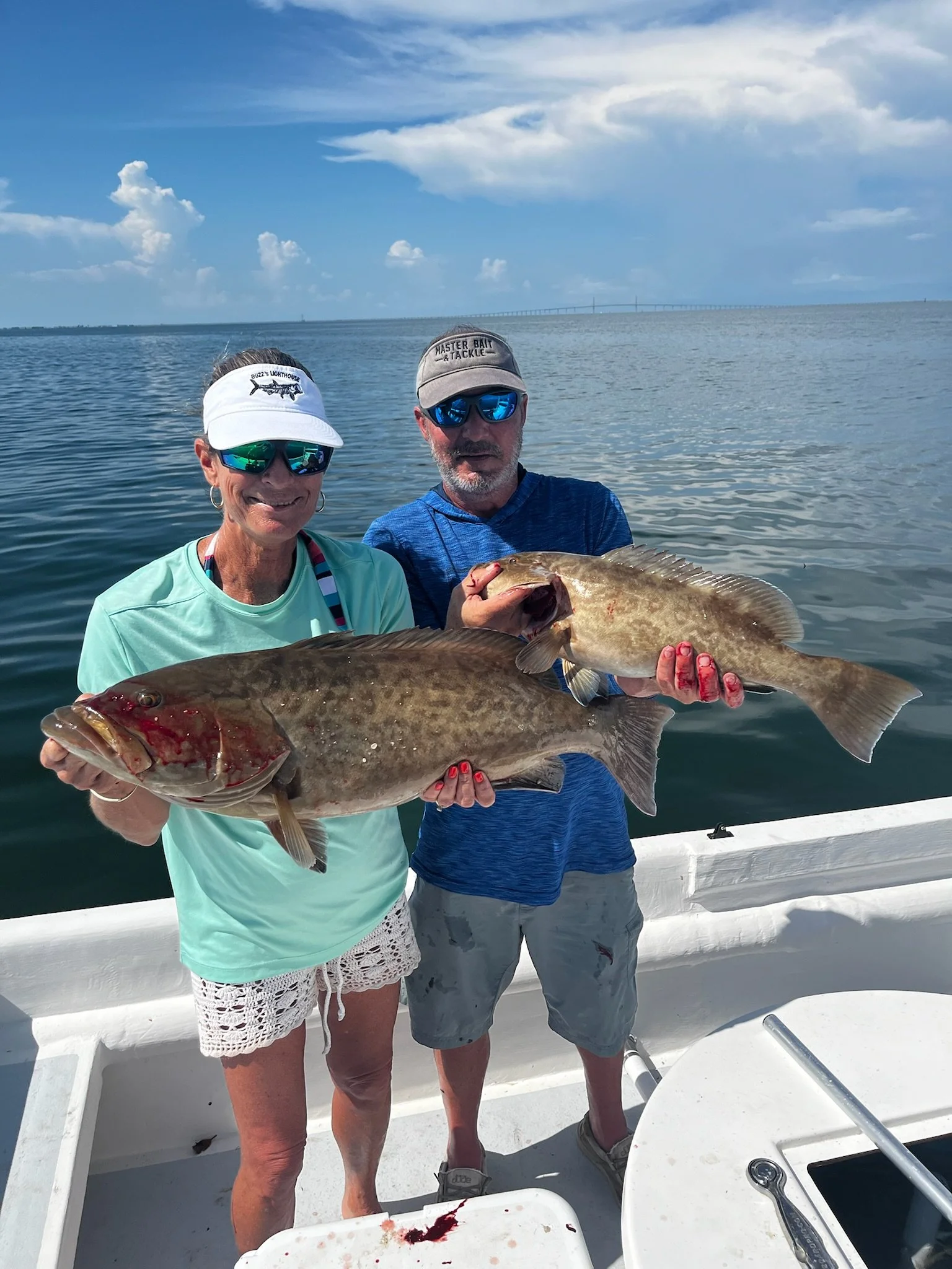 A man and woman on a boat holding a large fish, with the woman smiling and wearing a white visor and sunglasses, and the man wearing a cap labeled 'Master Bait & Tackle' and sunglasses. The boat is on water, and a distant bridge is visible in the background.