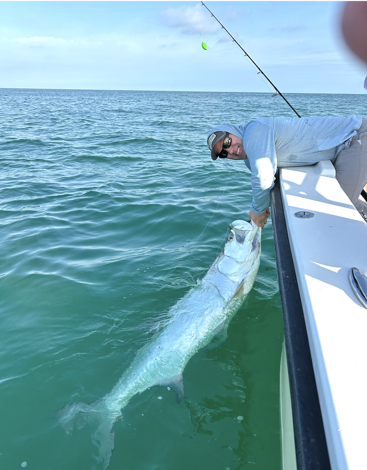 A man wearing sunglasses and a gray hoodie is on a boat holding a large fish, which he caught during a fishing trip on the open sea. The man is smiling and the water is a clear blue-green with gentle waves.