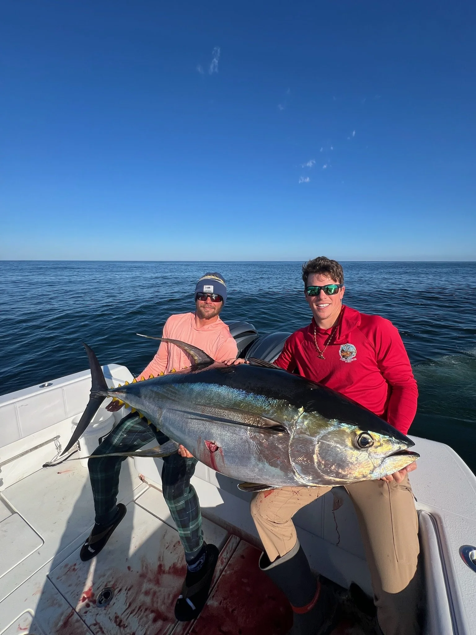 Two men on a boat holding a large fish with a blue ocean and clear sky in the background.