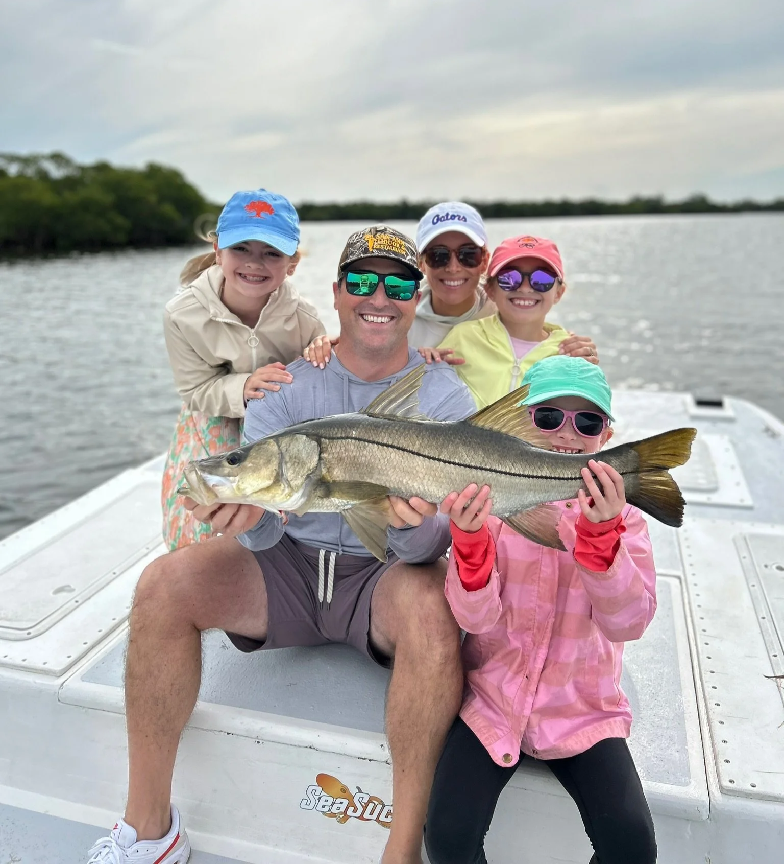 A group of five people, two children and three adults, on a boat holding a large fish during a fishing trip on a body of water with a background of trees and a cloudy sky.