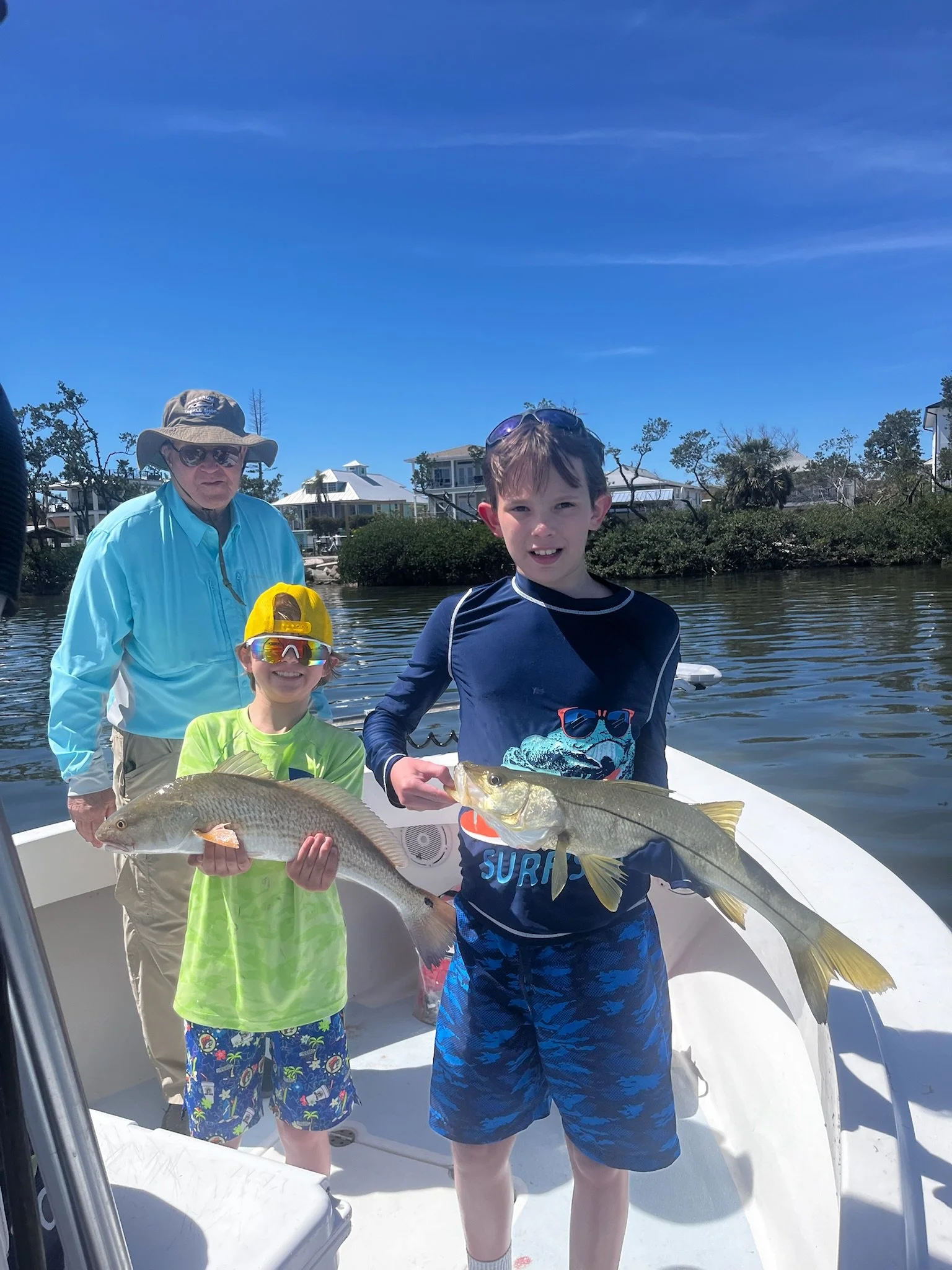 Two young boys and an older man on a boat holding fish with houses and trees in the background.