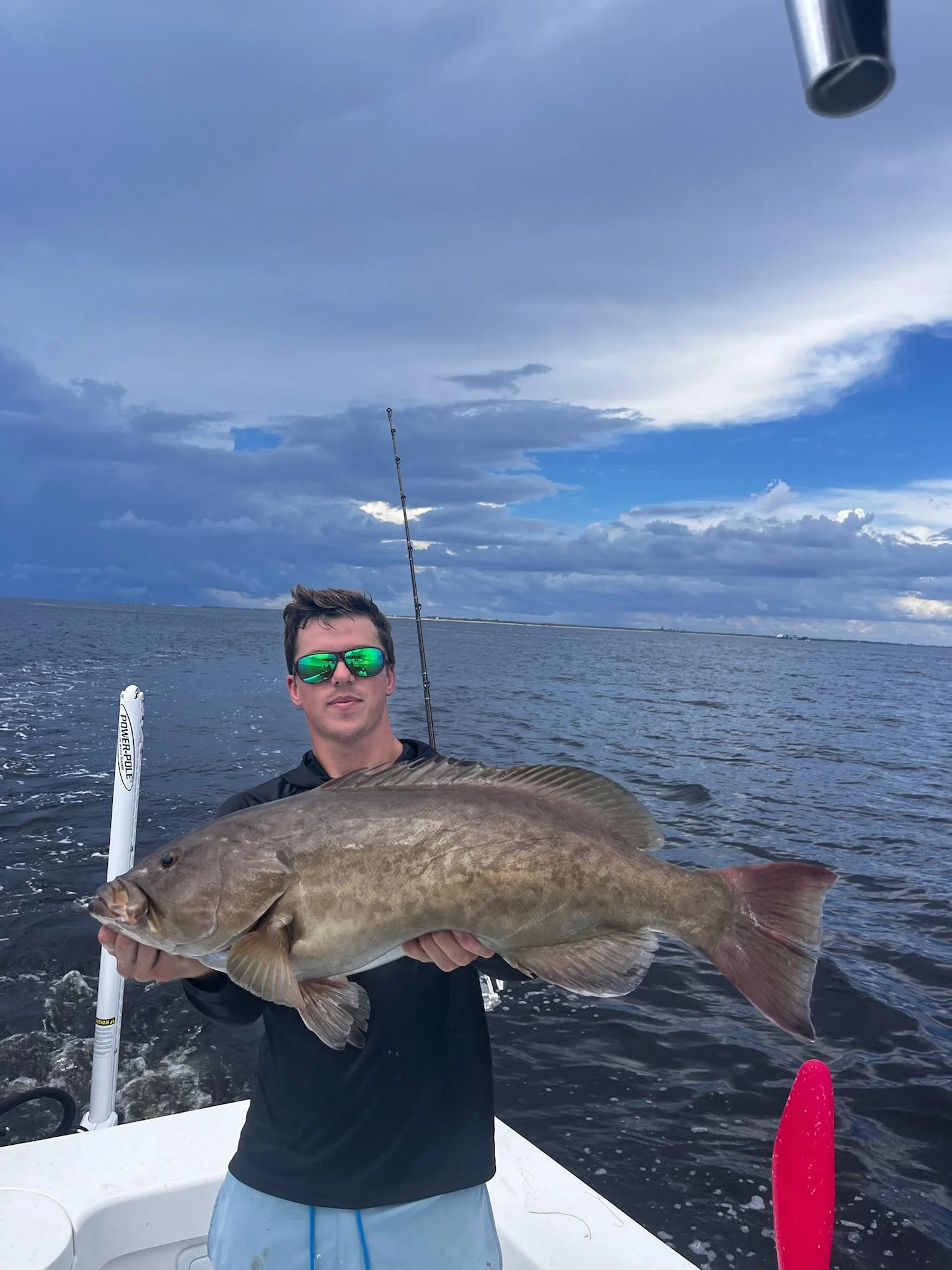 A man on a boat holding a large fish over water with a cloudy sky in the background.