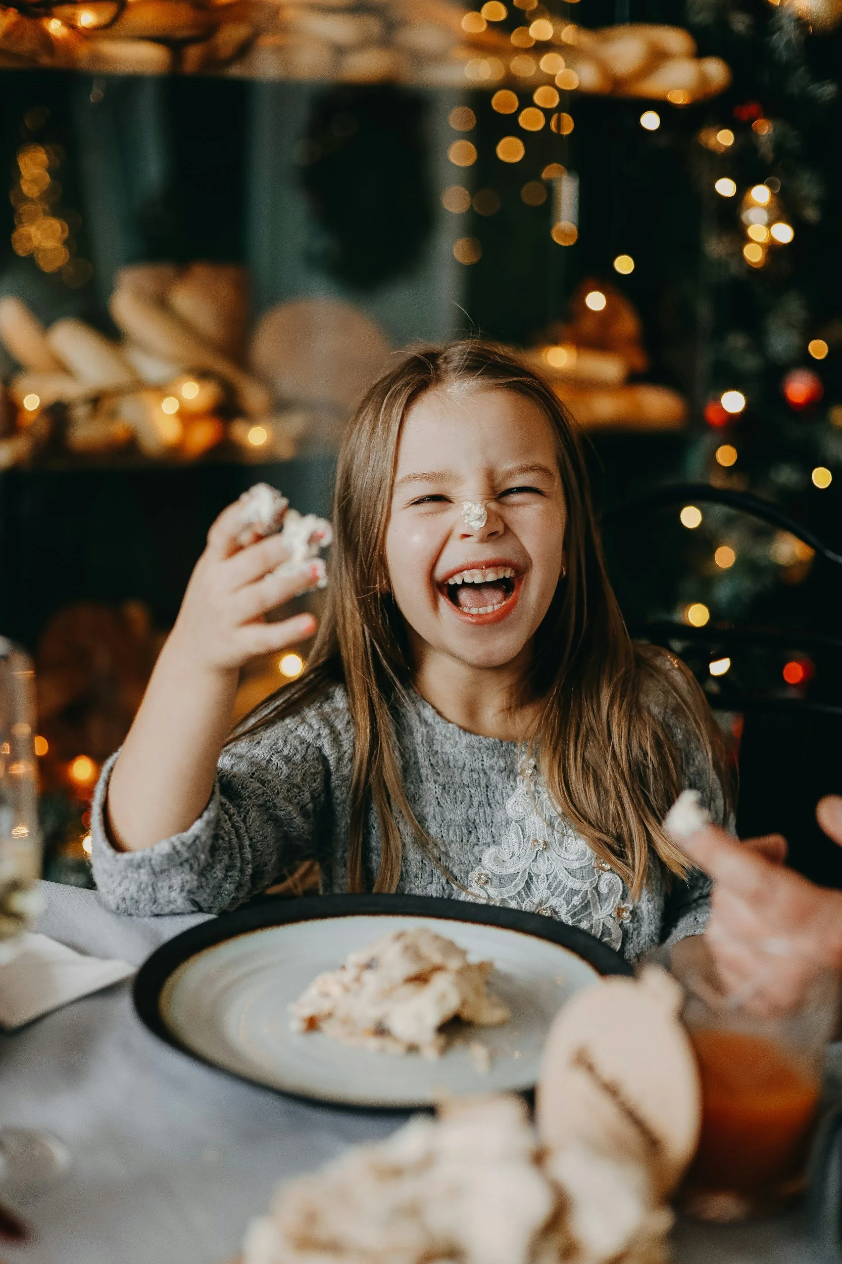 A young girl with long brown hair and a gray sweater is seated at a table, laughing and with a dollop of whipped cream on her nose. There are Christmas lights and decorations in the background. A plate with dessert and cookies is in front of her.