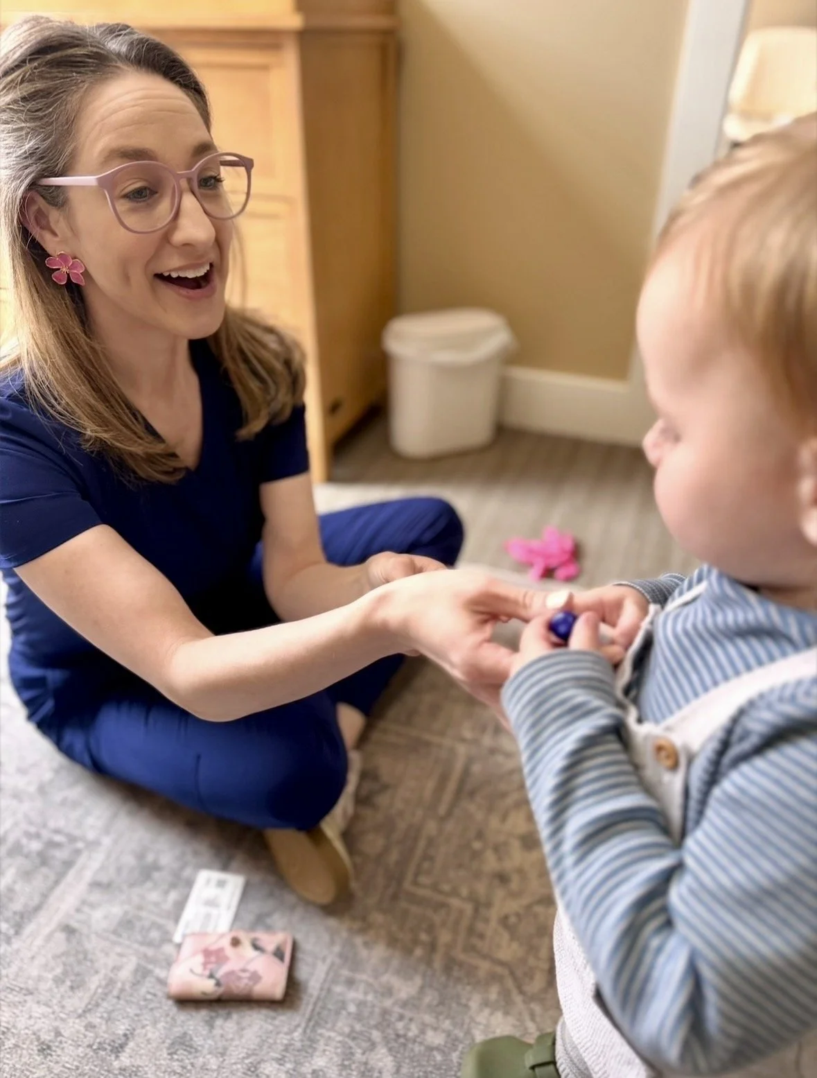 A woman with glasses and pink flower earrings is kneeling on the floor, smiling, and holding hands with a young boy in a striped shirt. There is a small pink toy on the floor behind them and a box of playing cards nearby.