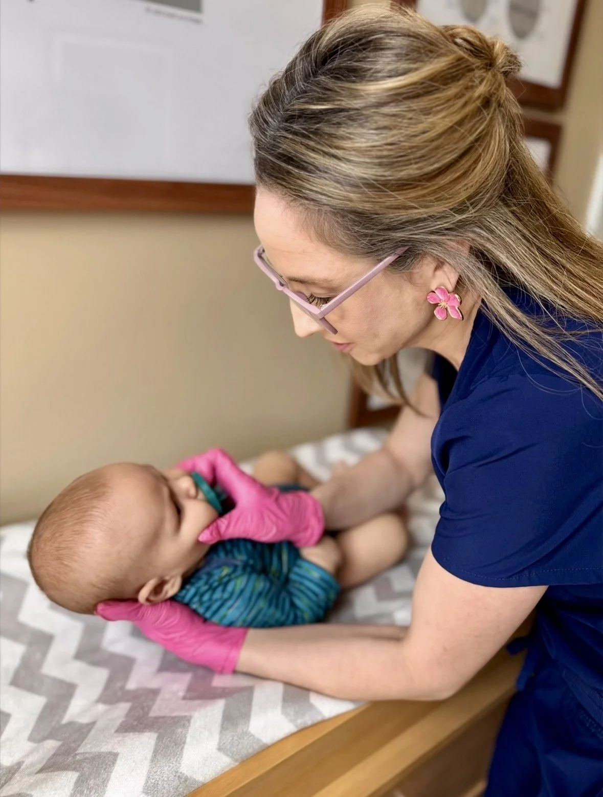 A woman in pink glasses and a dark blue shirt is holding a baby with pink gloves in a medical setting.