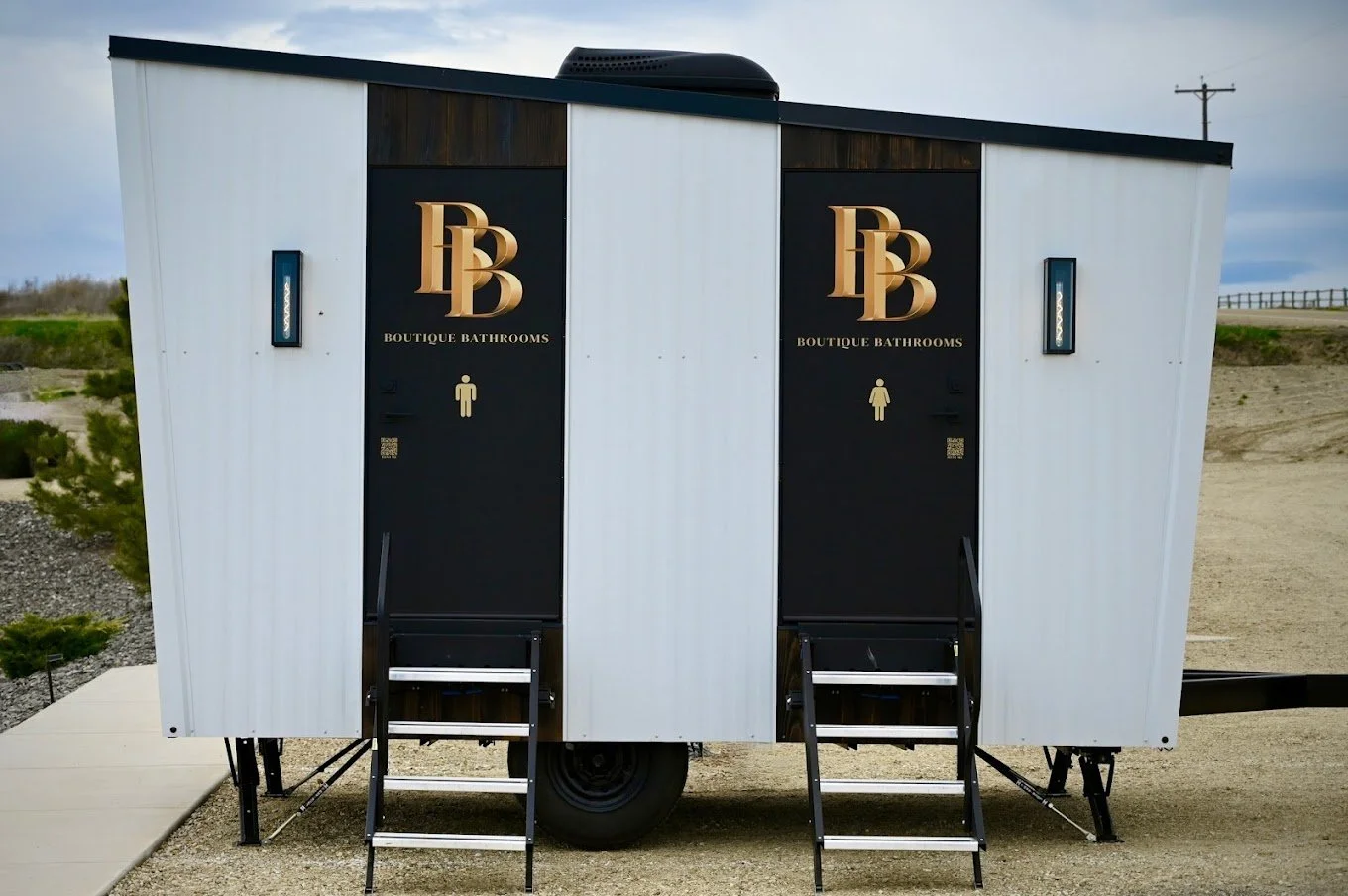 Portable building with double black doors labeled Bouique Bathrooms, two stairs leading to each door, and a logo on each door. The building is on wheels and situated outdoors on a gravel surface.