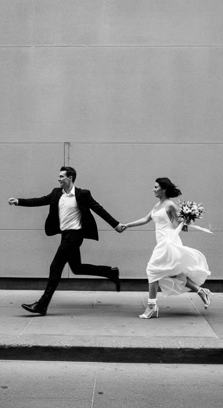 A black and white photo of a happy couple running hand in hand. The man is dressed in a suit and the woman in a wedding dress holding a bouquet of flowers.