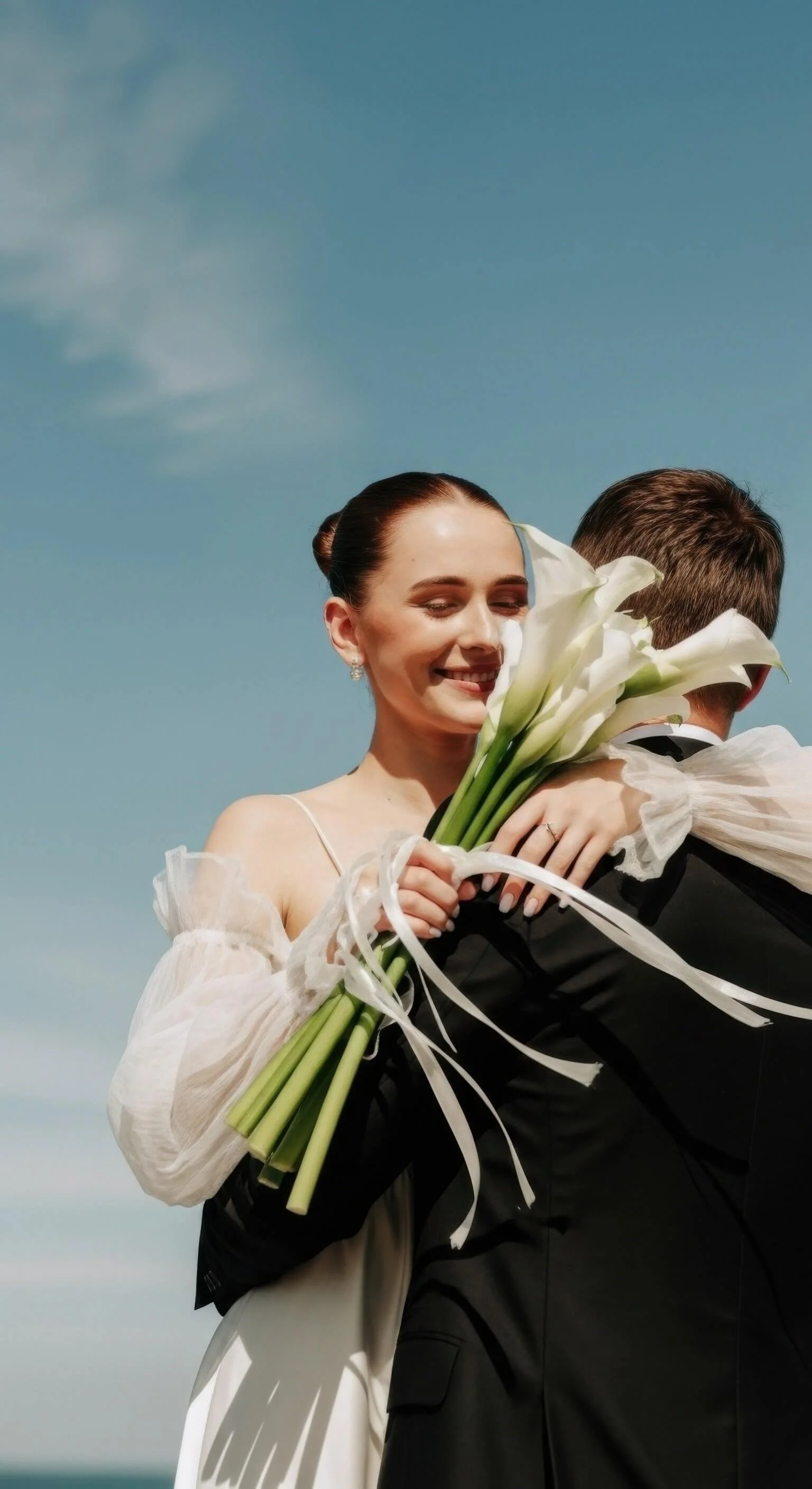 A woman in wedding attire is hugging a man in a tuxedo while holding a bouquet of white calla lilies against a blue sky backdrop.