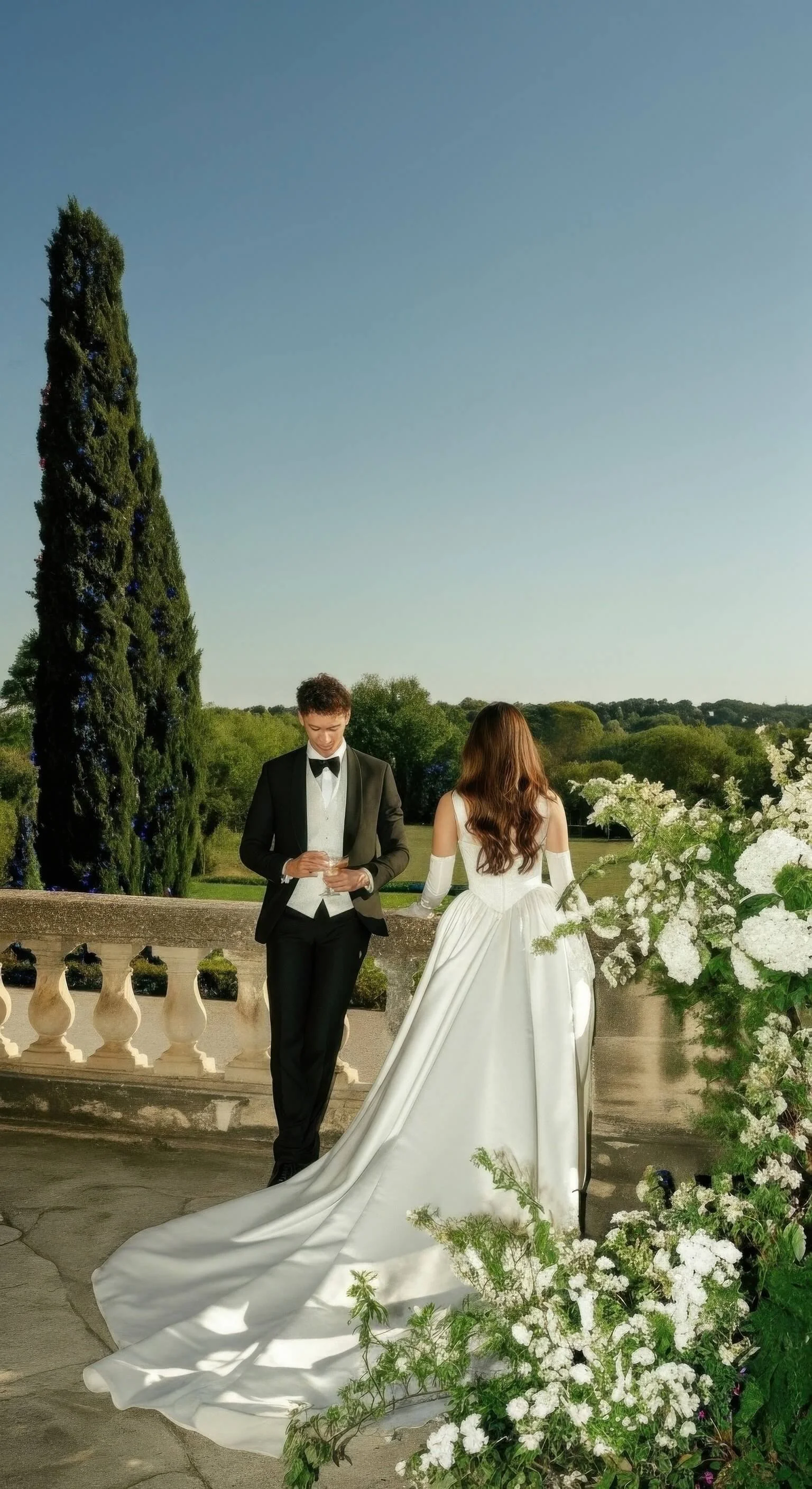 A groom in a black tuxedo and a bride in a white wedding gown stand on a terrace with floral arrangements, overlooking a green landscape with trees and a clear blue sky.