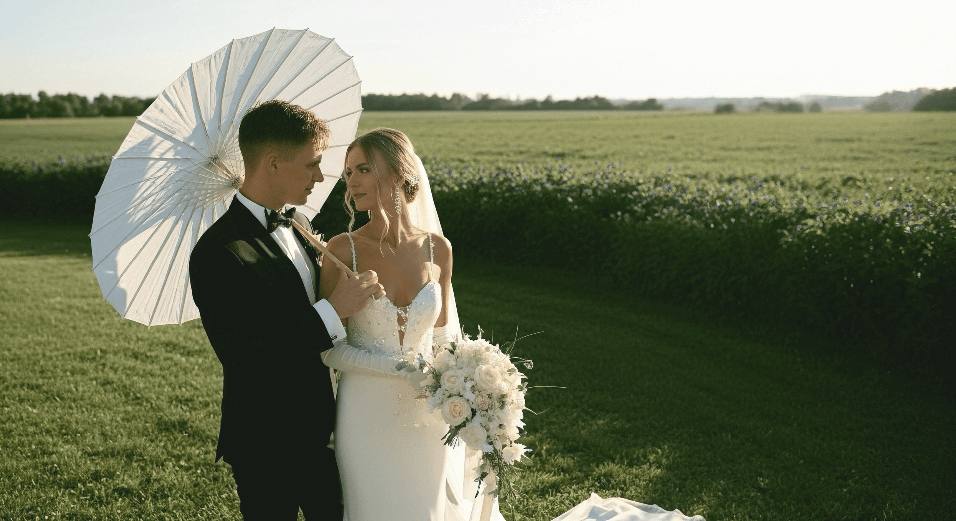 A bride and groom standing outdoors on a green field, holding a white umbrella. The groom is in a black tuxedo with a bow tie, and the bride is in a white wedding gown holding a bouquet of white roses. They are looking at each other under the umbrella with a distant landscape in the background.