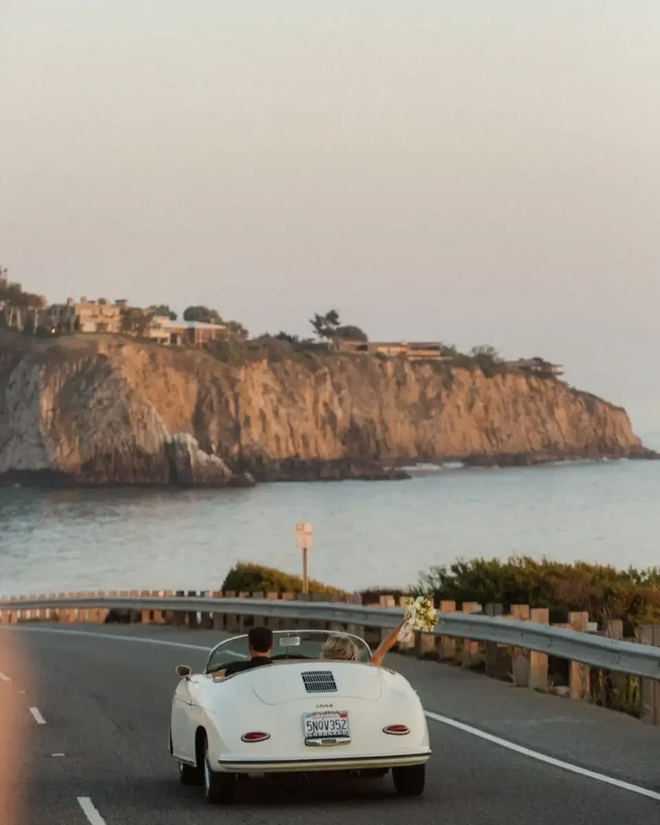 A white convertible car driving along a coastal road with a bride holding a bouquet of flowers. In the background, there are cliffs and the ocean at sunset.