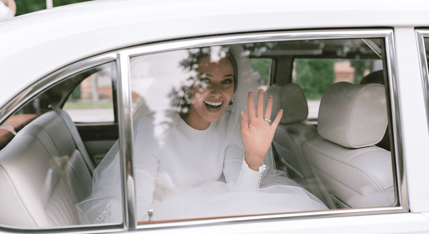 Bride sitting inside a wedding car, smiling and waving through the window.