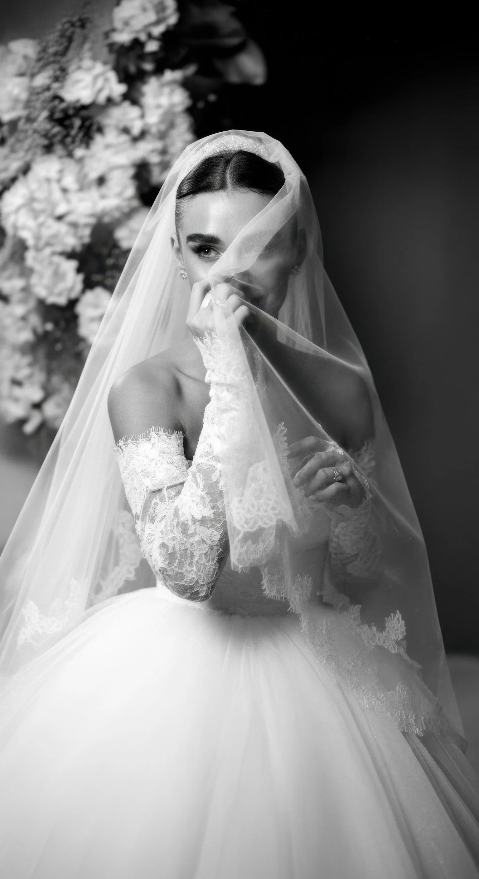 Black and white photo of a bride wearing a lace wedding dress and a veil, partially covering her face, with a background of flowers.