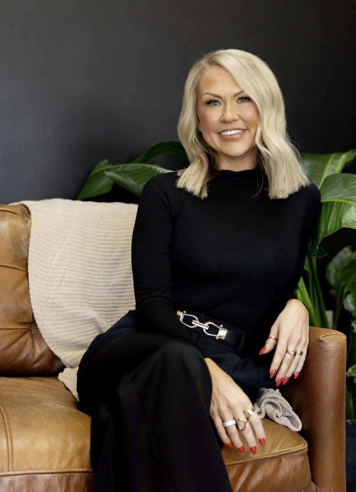 A woman with blonde hair, wearing a black long-sleeve top and black pants, is sitting on a tan leather couch with a cream blanket. She is smiling, showing her teeth, and has her right arm resting on her knee. She is wearing gold hoop earrings, rings on her fingers, and has red nail polish. Behind her are green plants and a dark gray wall.